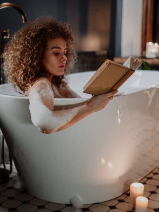 Self-care routine: a woman reading a book while taking a bubble bath with candles